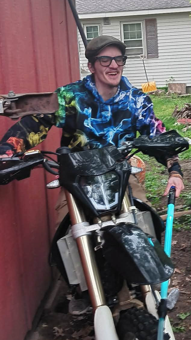 A man wearing glasses and a colorful patterned shirt sits on a dirt bike in an outdoor backyard setting next to a red shed.